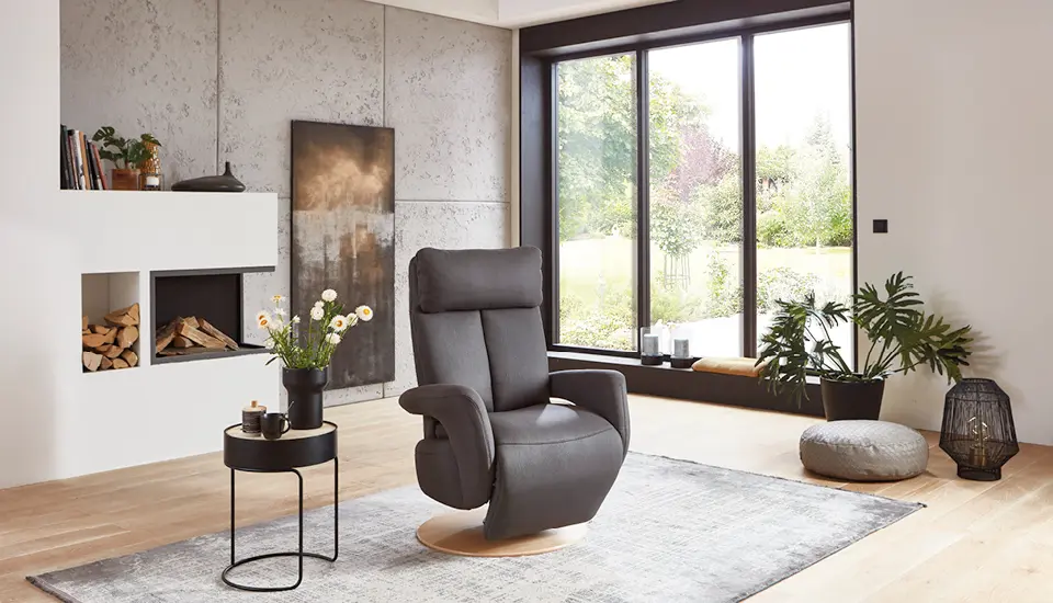 Komfortabler grauer Relaxsessel in modernem Wohnzimmer mit Kamin, großen Fenstern und stilvoller Dekoration, ideal für entspanntes Wohnen mit Blick in den Garten.